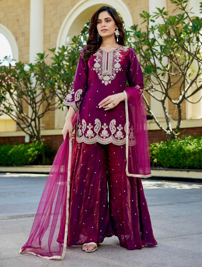 Woman wearing maroon ethnic suit with intricate gold embroidery and matching dupatta, standing outdoors