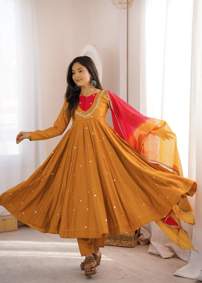 Front view of full three piece mustard yellow traditional festive outfit displaying an embroidered anarkali gown with reflective mirrors and trousers.